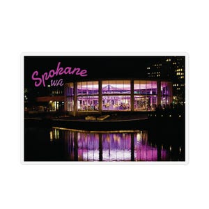 May include: A sticker featuring a nighttime view of a building in Spokane, WA, with the city name in pink script. The building has large windows and is reflected in the water below, with a purple glow.