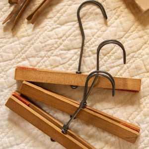 May include: Three wooden skirt hangers with dark metal hooks and orange felt lining on the interior clamping surfaces. The hangers feature a natural wood finish and are designed to grip garments securely between two parallel wooden bars.