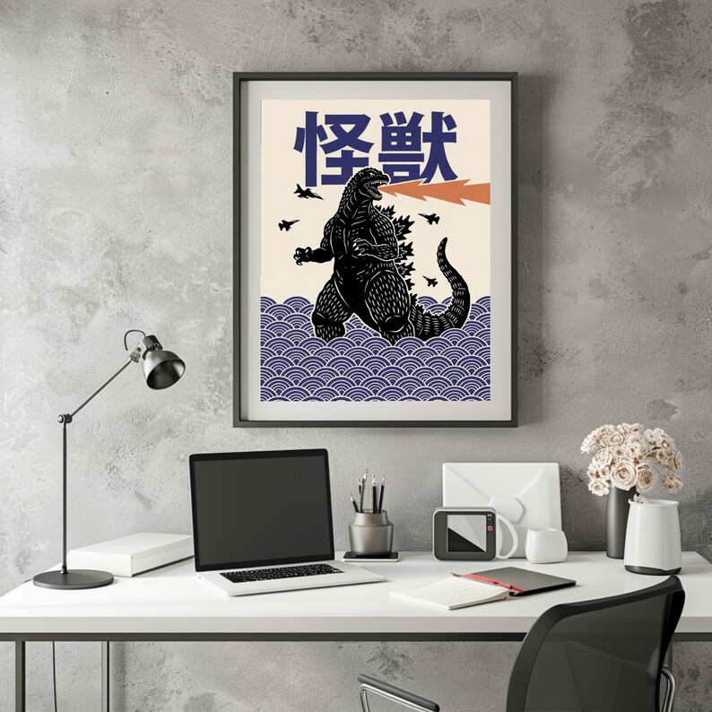 以下が含まれることがあります： 黒いゴジラのイラストが描かれた額入りのアートプリント。オレンジ色の火の息を吐き、クリーム色の背景に日本語のテキストと青い波模様が描かれています。アートワークは、ラップトップとデスクランプを備えた白い机の上にあります。
