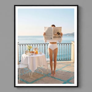 May include: A framed print of a person standing on a balcony, holding a newspaper with the headline "Prada Riviera". A small table with a white tablecloth is set with breakfast items. The background features the ocean and a clear blue sky.