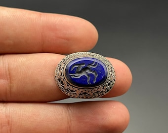 May include: A silver ring with a blue lapis lazuli stone set in a decorative silver bezel. The stone features a carved design of a stylized animal.