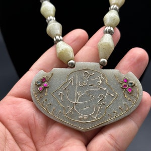May include: A jade necklace with a pendant featuring intricate gold calligraphy and pink gemstones. The pendant is shaped like a shield and has a silver backing. The necklace is made of white jade beads with silver accents.