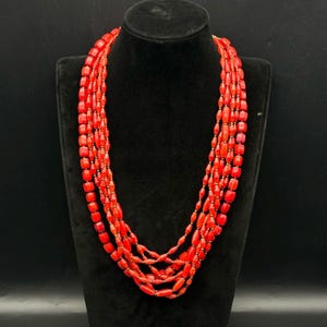 Può includere: Una collana multifilo realizzata con perline di corallo rosso vivo. La collana è composta da diversi fili di perline, e le perline sono tutte della stessa dimensione e forma.
