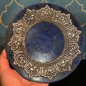 May include: A blue lapis lazuli dish with a silver rim and intricate floral designs. The dish is round and has a slightly raised centre.