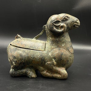 May include: A bronze sculpture of a ram with a hinged lid, possibly an incense burner or a container for small objects. The sculpture is in a kneeling position with its head turned to the side. The surface of the sculpture is covered in a green patina.