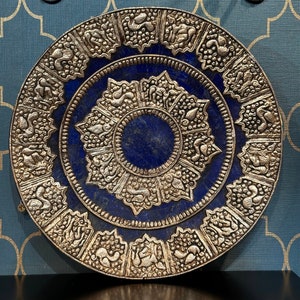 Op de afbeelding: Een ronde, zilverkleurige decoratieve plaat met een blauw lapis lazuli centrum. De plaat heeft ingewikkelde, gegraveerde ontwerpen rond de rand en in het midden.