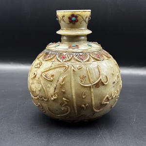 May include: A white jade bottle with intricate carvings and inlaid turquoise and red stones. The bottle has a round body and a narrow neck with a flared top. The bottle is decorated with floral motifs and Arabic calligraphy.