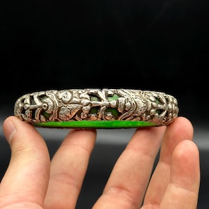 Puede incluir: Un brazalete de plata con intrincados diseños florales y animales, con una incrustación de jade verde.