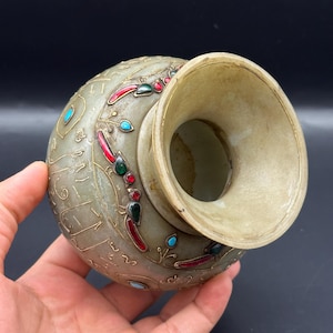 May include: A small, round, green jade vase with intricate gold and red enamel designs and turquoise and red gemstones. The vase has a wide opening and a slightly flared lip.