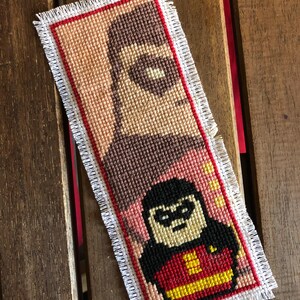 May include: Handmade cross-stitch bookmark featuring a superhero design. The bookmark has a red border and a white fringe. The design depicts a character in a black mask and red and yellow costume. The background is a mix of pink and brown.