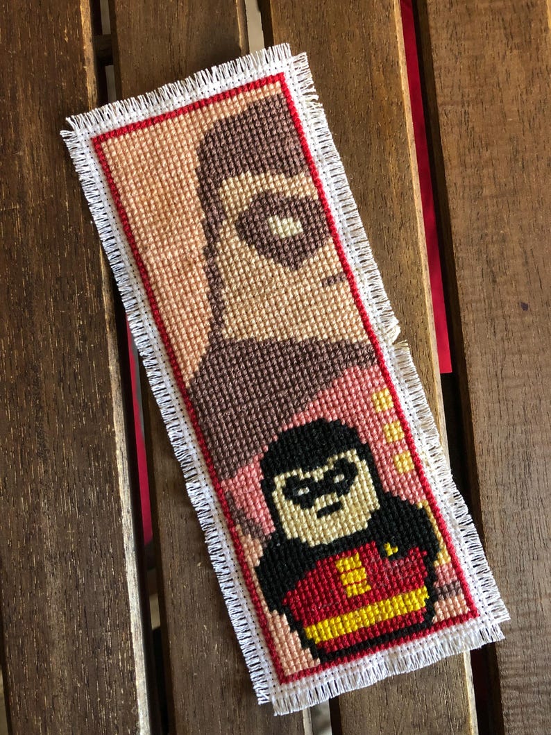 May include: Handmade cross-stitch bookmark featuring a superhero design. The bookmark has a red border and a white fringe. The design depicts a character in a black mask and red and yellow costume. The background is a mix of pink and brown.