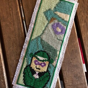 May include: Cross-stitched bookmark featuring a green and purple character design. The bookmark has a white fringe border and a green border. The design is on a light green background. The bookmark is rectangular and appears to be handmade.