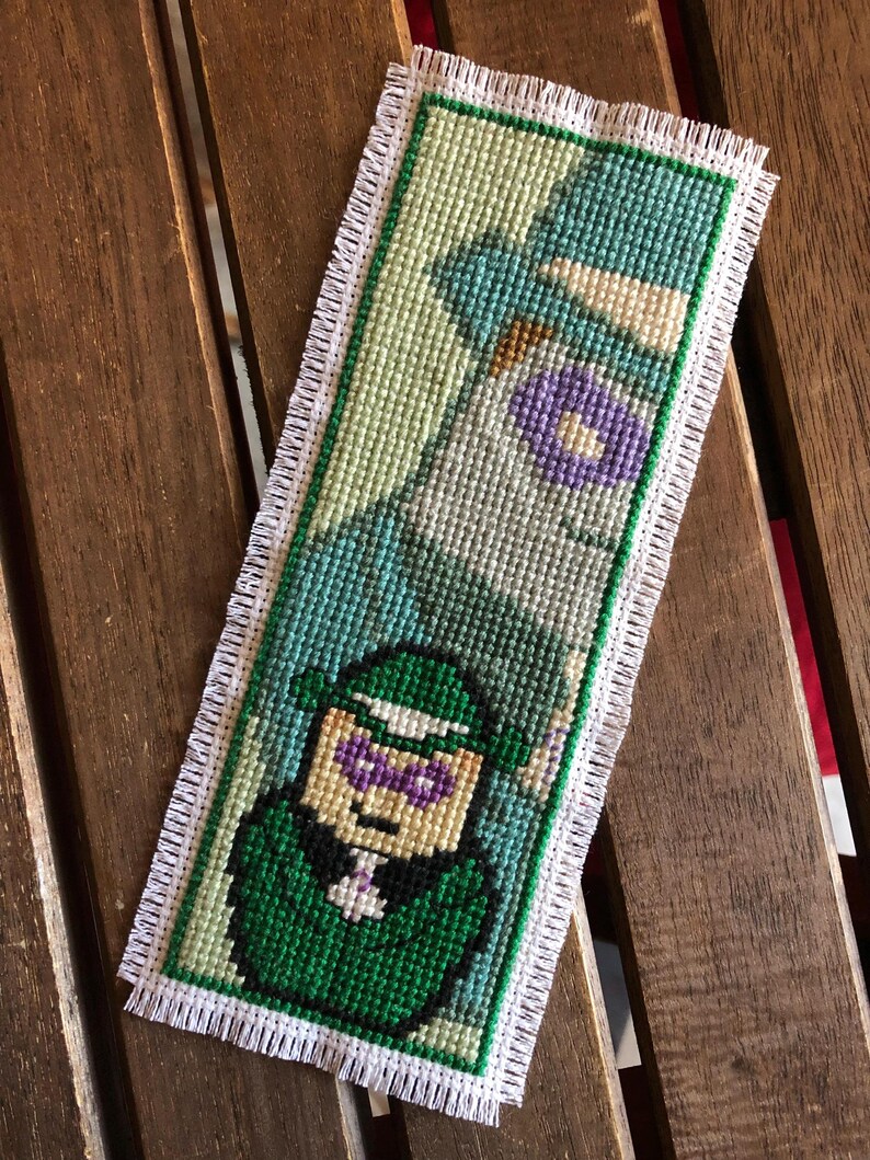 May include: Cross-stitched bookmark featuring a green and purple character design. The bookmark has a white fringe border and a green border. The design is on a light green background. The bookmark is rectangular and appears to be handmade.