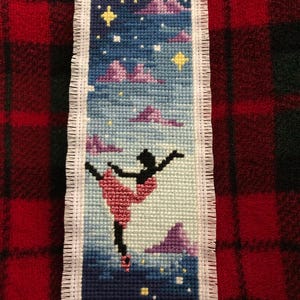 May include: Cross-stitch bookmark featuring a ballerina in a pink tutu, set against a night sky with stars and clouds. The bookmark has a white fringed border and is made with various shades of blue, pink, and yellow.