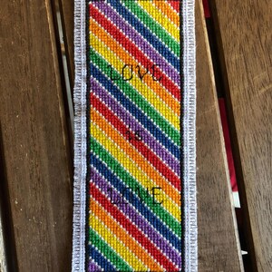 Può includere: Segnalibro fatto a mano con un motivo arcobaleno a strisce diagonali rosse, arancioni, gialle, verdi, blu e viola. Le parole "LOVE" e "ME" sono ricamate in bianco. I bordi sono rifiniti con frange bianche.