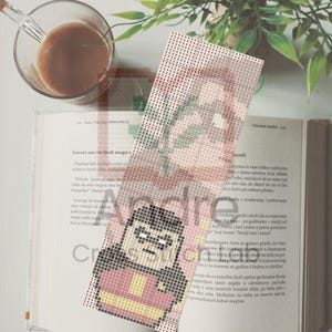 May include: A handmade cross-stitch bookmark featuring a cartoon character in a red and black outfit. The bookmark is placed on an open book with text. A cup of coffee sits in the upper left corner.