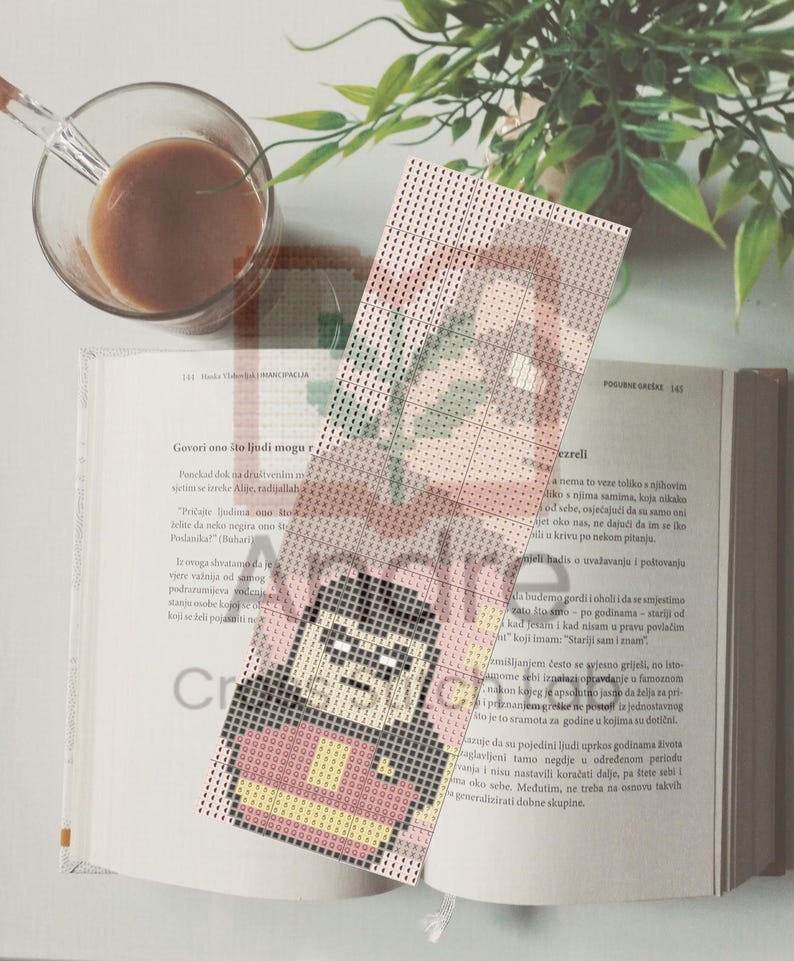 May include: A handmade cross-stitch bookmark featuring a cartoon character in a red and black outfit. The bookmark is placed on an open book with text. A cup of coffee sits in the upper left corner.