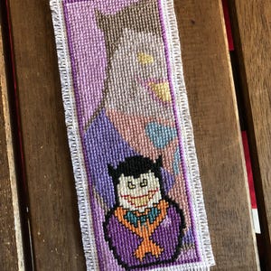 May include: Handmade cross-stitch bookmark featuring a cartoon vampire design. The bookmark has a purple background with a white fringe border. The vampire is depicted with a black cape, purple suit, and orange bow tie. The bookmark is rectangular.