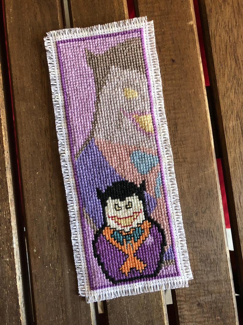 May include: Handmade cross-stitch bookmark featuring a cartoon vampire design. The bookmark has a purple background with a white fringe border. The vampire is depicted with a black cape, purple suit, and orange bow tie. The bookmark is rectangular.