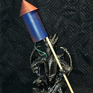 Puede incluir: Un cohete de fuegos artificiales azul y naranja es sostenido por una estatua de dragón oscura y detallada. El cohete tiene un cuerpo azul y una parte superior en forma de cono naranja. El dragón está intrincadamente esculpido con alas y garras.