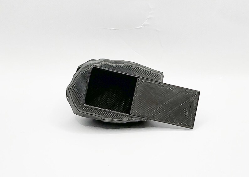 Puede incluir: Una caja negra en forma de roca, impresa en 3D, con una tapa con bisagras. La tapa est&aacute; abierta, revelando el interior vac&iacute;o de la caja.