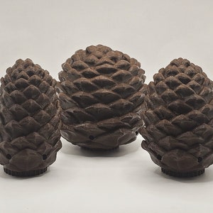 以下が含まれることがあります： 茶色の松ぼっくり形のオーナメントが3つ。松ぼっくりは質感のある素材で作られており、リアルな外観をしています。