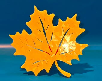 May include: A vibrant, orange maple leaf decoration with a detailed, geometric design. The leaf has a stem and is set against a deep blue background, casting a shadow. The leaf's veins are subtly marked with red.