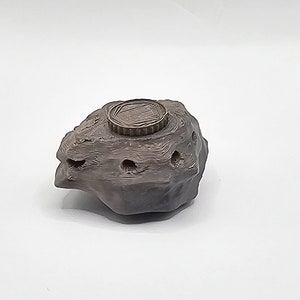 Puede incluir: Una peque&ntilde;a roca de cer&aacute;mica gris oscuro con una parte superior redonda, texturizada y de color bronce. La roca tiene varios agujeros peque&ntilde;os en el lateral.