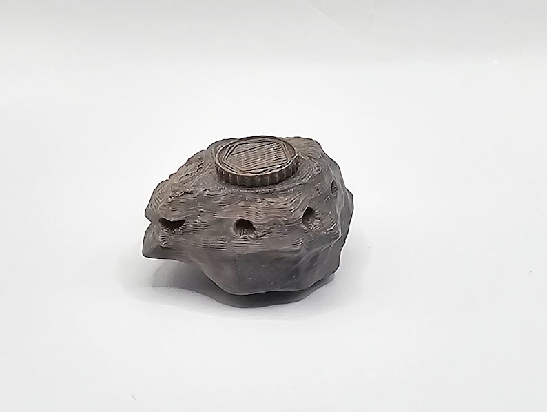 Puede incluir: Una peque&ntilde;a roca de cer&aacute;mica gris oscuro con una parte superior redonda, texturizada y de color bronce. La roca tiene varios agujeros peque&ntilde;os en el lateral.