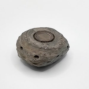 Puede incluir: Una peque&ntilde;a roca gris impresa en 3D con una parte superior redonda, texturizada y marr&oacute;n. La parte superior tiene una abertura hexagonal con una tapa marr&oacute;n texturizada y elevada.