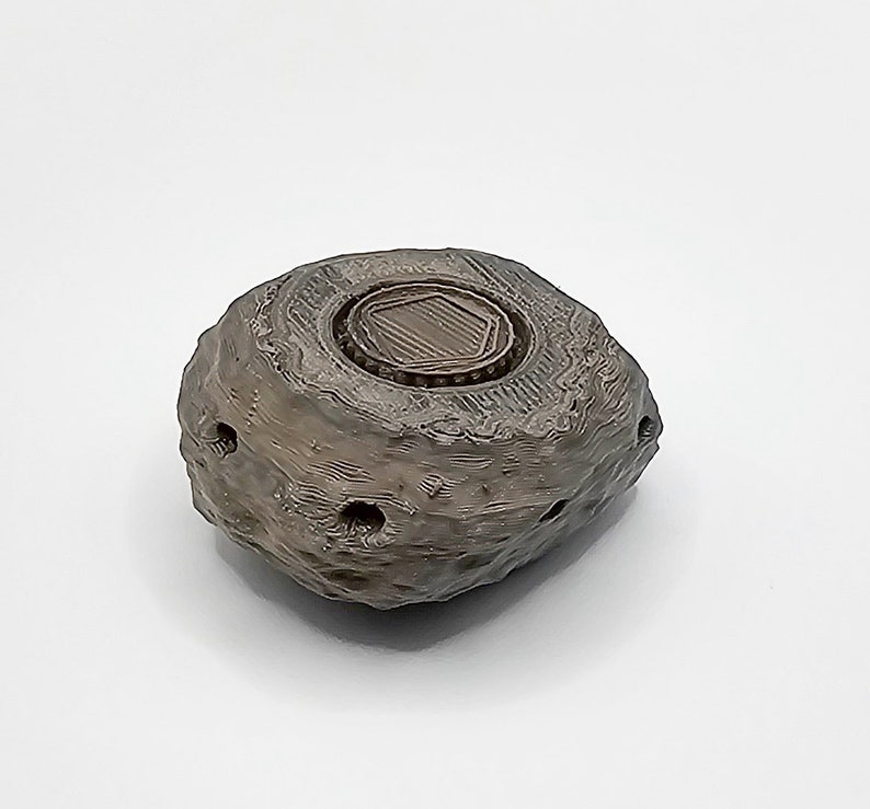 Puede incluir: Una peque&ntilde;a roca gris impresa en 3D con una parte superior redonda, texturizada y marr&oacute;n. La parte superior tiene una abertura hexagonal con una tapa marr&oacute;n texturizada y elevada.