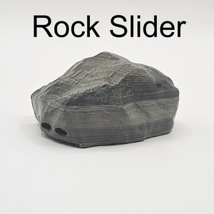 Puede incluir: Una roca gris impresa en 3D con una superficie texturizada. La roca tiene una base plana y est&aacute; dise&ntilde;ada para deslizarse sobre una superficie.