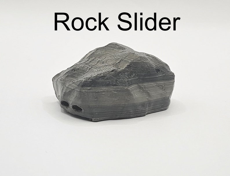 Puede incluir: Una roca gris impresa en 3D con una superficie texturizada. La roca tiene una base plana y est&aacute; dise&ntilde;ada para deslizarse sobre una superficie.