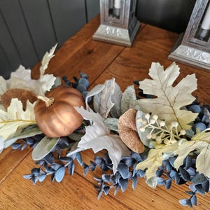 May include: A fall-themed decorative garland featuring a metallic copper pumpkin, cream-colored leaves, and blue-green foliage. The arrangement is set on a wooden surface, creating a warm, autumnal aesthetic. Decorative lanterns are in the background.