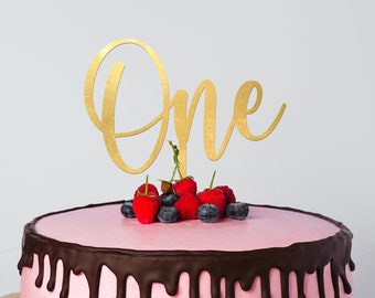 May include: A pink cake with chocolate frosting and a gold 'One' topper. The cake is decorated with fresh raspberries and blueberries.