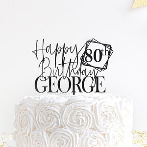 May include: Black acrylic cake topper with the text "Happy Birthday George" and "80th" in a square frame. The topper is on a white cake with white frosting.