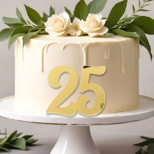 May include: A white cake with gold drip icing and a gold number 25 topper. The cake is decorated with white roses and green leaves.