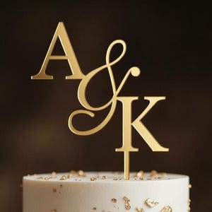 May include: A gold-colored cake topper with the letters "A & K" on top of a white cake. The cake is decorated with gold leaf accents. The background is a blurred, dark brown.