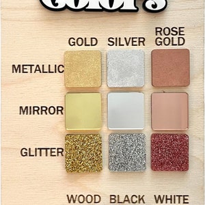 May include: A wooden board with the word "Colors" written in black and white lettering. The board displays a variety of colour swatches in different finishes, including metallic, mirror, glitter, wood, black, and white.