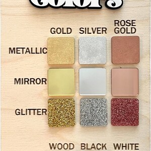 May include: A wooden board with the word "Colors" written in black and white lettering. The board displays a variety of color samples, including metallic gold, silver, rose gold, mirror gold, silver, rose gold, glitter gold, silver, rose gold, wood, black, and white.