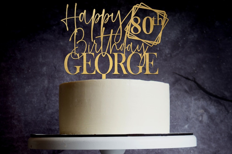 May include: Gold cake topper with the text "Happy 80th Birthday GEORGE" in a modern calligraphy style. The number 80 is framed in a geometric design.