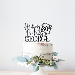 May include: Black cake topper with the text "Happy Birthday GEORGE" and "80th" in a square frame. The topper is on a white cake with a light brown frosting.