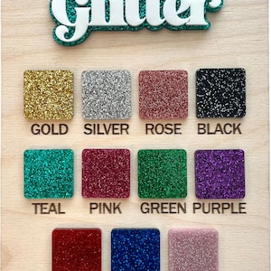 May include: A wooden board with the word "Glitter" in large white letters with a teal outline. The board displays a colour chart of 12 different glitter colours, including gold, silver, rose, black, teal, pink, green, purple, red, navy, and blush.