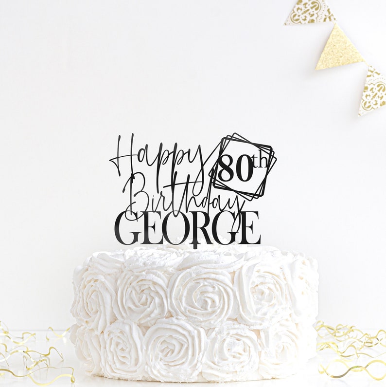 May include: Black acrylic cake topper with the text "Happy Birthday George" and "80th" in a square frame. The topper is on a white cake with white frosting.