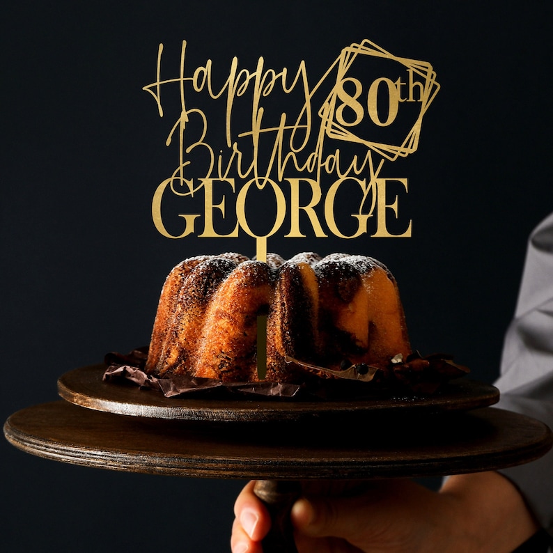 May include: Gold cake topper with the text "Happy 80th Birthday GEORGE" in a modern calligraphy font. The topper is on a cake with white powdered sugar.