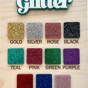 May include: A wooden board with the word "Glitter" in large white letters with a teal outline. The board displays a color chart of 12 different glitter colors, including gold, silver, rose, black, teal, pink, green, purple, red, navy, and blush.