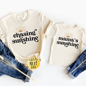 May include: Two beige t-shirts with black text and a gold sun graphic. The larger shirt says "chasing sunshine" and the smaller shirt says "mama's sunshine".