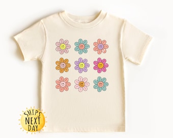 May include: A cream colored t-shirt with a pattern of nine colorful flowers with smiley faces. The flowers are in shades of pink, blue, yellow, and orange.