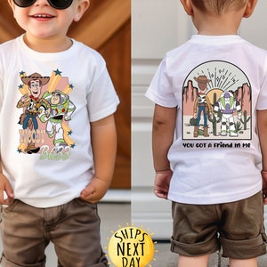 K&ouml;nnte beinhalten: Wei&szlig;es T-Shirt mit einem Cartoon-Bild von Woody und Buzz Lightyear aus Toy Story. Die Vorderseite des Shirts tr&auml;gt den Schriftzug "Woody Buzz". Die R&uuml;ckseite des Shirts zeigt ein Cartoon-Bild von Woody und Buzz Lightyear in einer W&uuml;stenlandschaft mit dem Text "You got a friend in me".