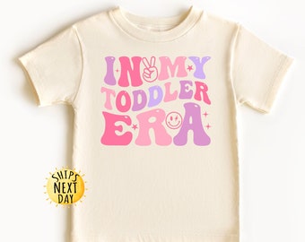 May include: A cream coloured t-shirt with the text "In My Toddler Era" in pink, purple, and white with a peace sign and a smiley face.